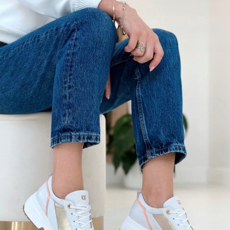 Kobieta w jeansach i białej bluzie siedzi na złotym stołku, prezentując białe sneakersy Ulmani z pomarańczowymi i złotymi detalami.